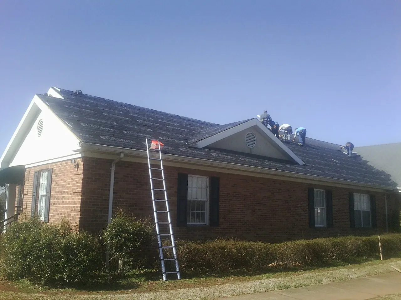 Crew tearing shingles off a brick ranch roof with a ladder set — Emergency Roof Repair work by our crew in Mitchellville