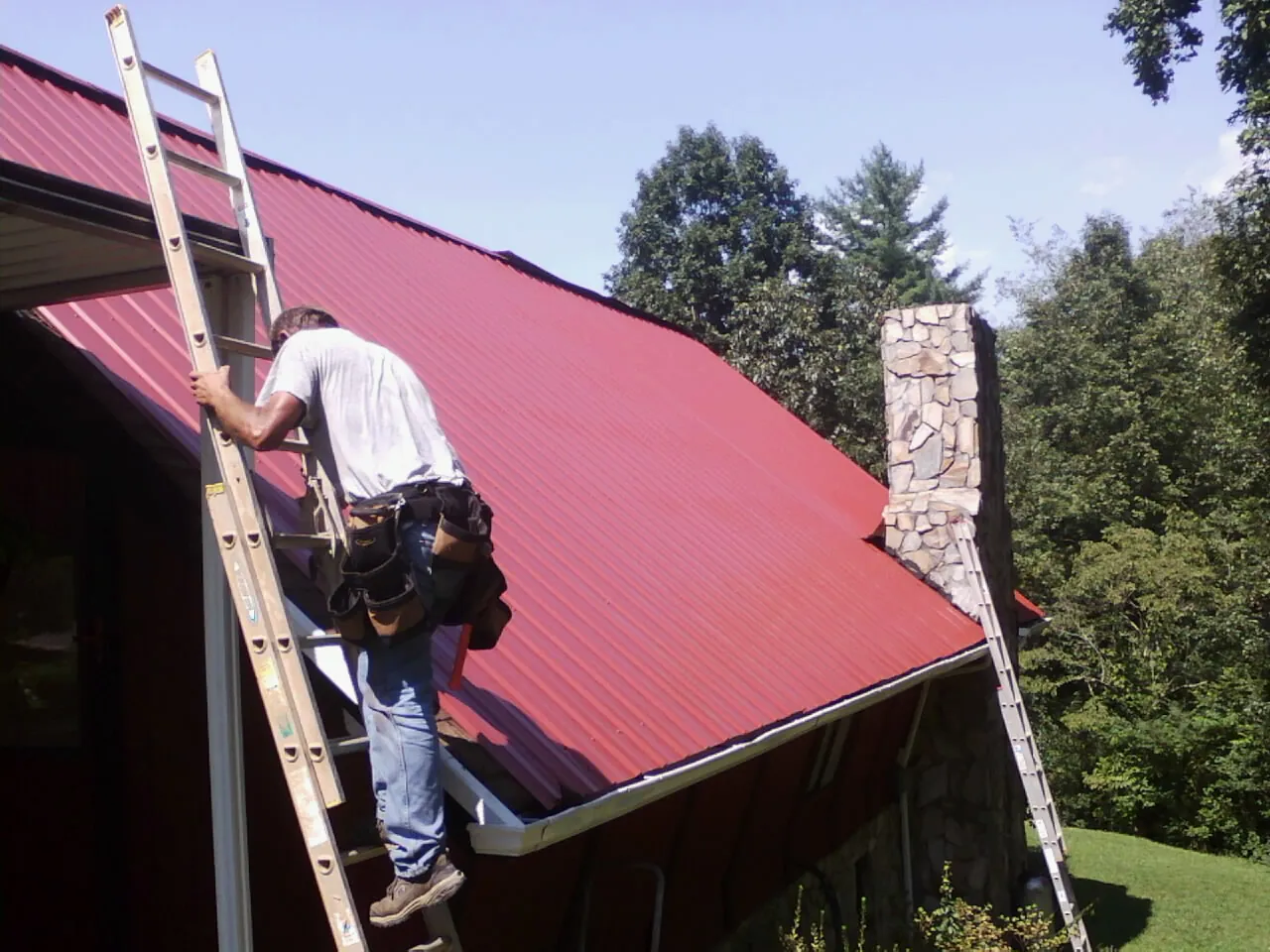 Roofer on a ladder beside a red metal roof and stone chimney — Soffit & Fascia Repair by Roofing Brothers in Mitchellville